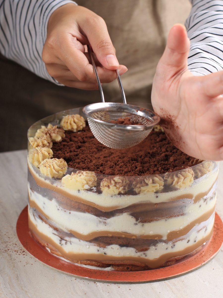 Tort tiramisu, otoczony folią wspomagającą, podczas nasypywanie kakao na wierzch deseru.